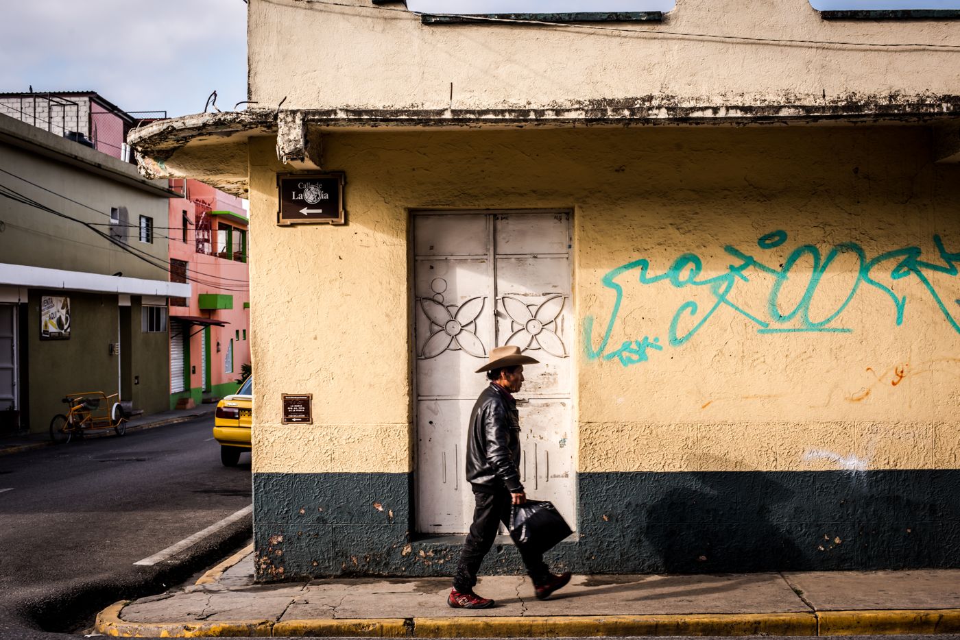 A man in a cowboy hat walking alone past a yellow building in Oaxaca